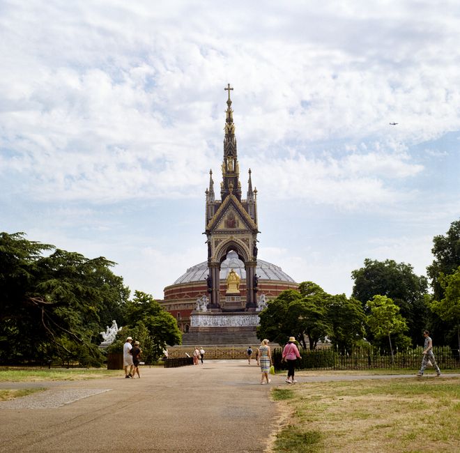 A wide pathway through Queen's Park with a sandy surface showing patches of puddles and dampness, flanked by leafless trees, leading towards a historic monument with a tall spire in the background. Th