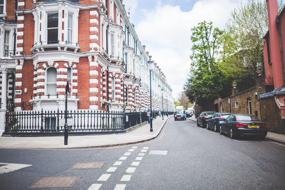 A quiet residential street in Queen's Park NW6 with a row of historic Victorian-style terraced houses featuring red and white brick facades, decorative white window frames, and ornate cornices. The pavement is clean, with a black wrought-iron fence enclosing a small front garden for one property. Several modern cars are parked along the curb on the right side of the street, including a silver sedan and a black vehicle, while the street itself has clear white dashed lane markings. Tall, leafy green trees line the sidewalk, providing a touch of nature and shade. The scene is well-lit with natural daylight, highlighting the tidy, well-maintained appearance of the neighborhood. Queen's park Cleaners offers comprehensive cleaning and deep cleaning services, ensuring homes maintain high standards of hygiene and sanitisation, which is reflected in the pristine condition of this residential street.