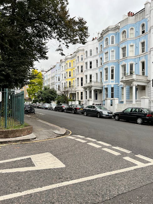 A street scene in Queens Park featuring a row of Victorian-style residential buildings painted in pastel shades of blue, white, and yellow. The buildings have ornate architectural details such as decorative moldings and bay windows. The street is lined with parked cars of various models and colors, including black, silver, and white. On the left, a sidewalk with a metal fence surrounds a green area with trees, some of which have dense foliage extending over the street. The pavement appears clean and well-maintained, with a pedestrian crossing visible in the foreground. The overall scene showcases a typical residential street in Queens Park, highlighting the aesthetic urban environment that Queen's Park Cleaners services through professional cleaning and cleaning maintenance for both residential buildings and public spaces.