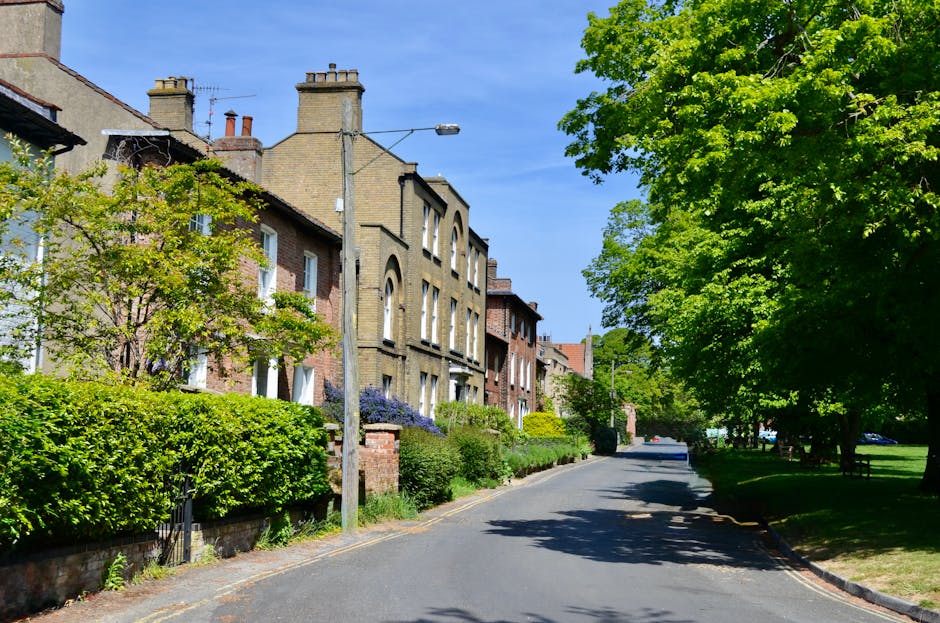 A quiet residential street in Queen's Park NW6 with a row of historic Victorian-style terraced houses featuring red and white brick facades, decorative white window frames, and ornate cornices. The pavement is clean, with a black wrought-iron fence enclosing a small front garden for one property. Several modern cars are parked along the curb on the right side of the street, including a silver sedan and a black vehicle, while the street itself has clear white dashed lane markings. Tall, leafy green trees line the sidewalk, providing a touch of nature and shade. The scene is well-lit with natural daylight, highlighting the tidy, well-maintained appearance of the neighborhood. Queen's park Cleaners offers comprehensive cleaning and deep cleaning services, ensuring homes maintain high standards of hygiene and sanitisation, which is reflected in the pristine condition of this residential street.